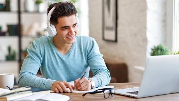 A learner wearing headphones takes notes during an online tutoring session at his laptop