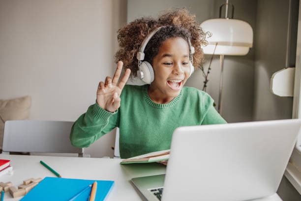 A young student with headphones waves hello to her tutor during a video lesson